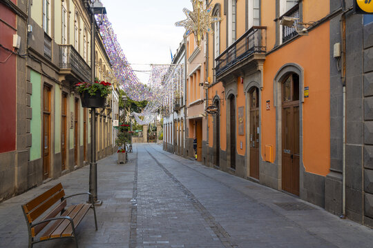 Arucas, Spain - 27 December 2025: View of the historic street in Arucas with colorful colonial-style buildings, a wooden bench, and festive holiday decorations hanging overhead.