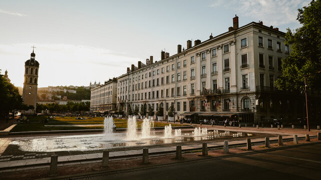Lyon, France - 10 May 2022: View of the Place Bellecour square with its fountain, historic buildings, and church tower in the background during a warm sunset.