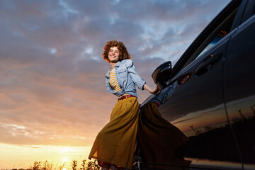 Obraz premium Cheerful woman outdoors, wearing a denim jacket and yellow skirt, stepping from a car at sunset, laughter in her eyes, warm light, relaxed mood, natural colors