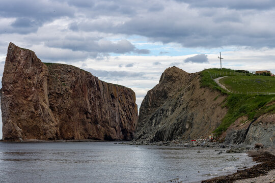 Gaspesie, Quebec, 15 July 2026 : Large rock formation stands in ocean water next to a cliff with a cross on a grassy hill.