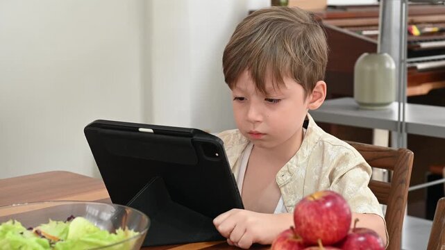Young boy focused on tablet at dining table with salad bowl and apple fruit indoor digital tablet at kitchen table suggesting quiet concentration and casual morning routine
