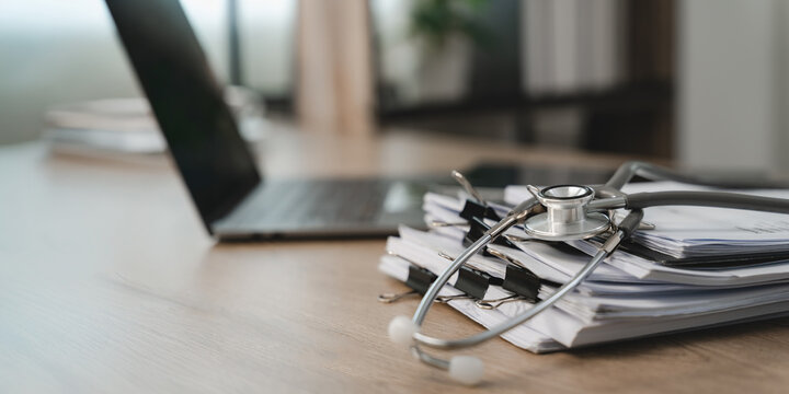 Stethoscope resting on stacked paper files beside a laptop at an office desk, showing healthcare documentation, medical record organization and a clinical paperwork management workflow.