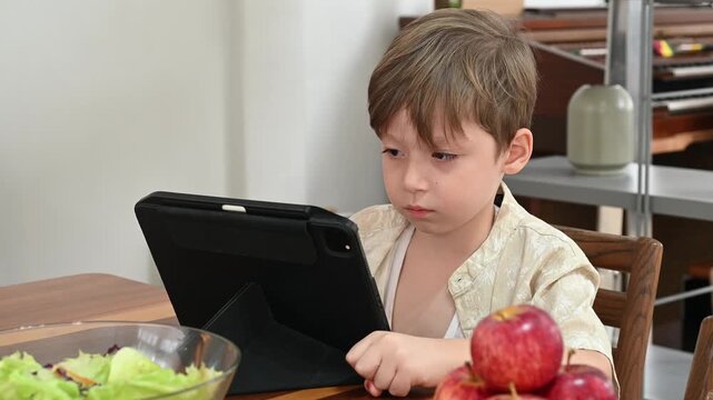 Young boy focused on tablet at kitchen table with salad bowl and apples nearby, calm breakfast scene of child and fruit in natural light as screen learning and quiet concentration