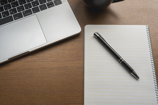 A laptop keyboard sits beside a spiral notebook and pen on a wooden desk, with a mug nearby.