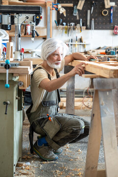 Passionate craftsman attentively checking surface of antique wooden door at worktable in workshop, man carpenter examining wood condition, scrutiny during reconstruction in woodworking studio.