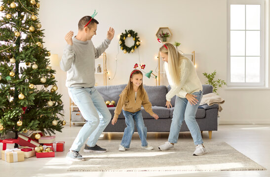 Christmas family having fun dancing with child girl at home. Parents and daughter dance near decorated festive tree with gifts, sharing warm winter holiday celebration. New year joy concept.