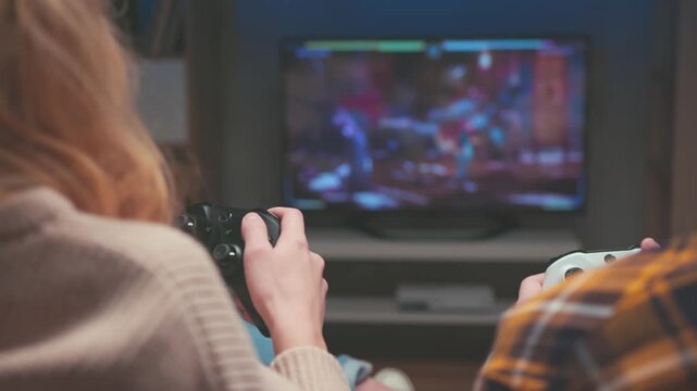Unrecognizable Couple Competing with White and Black Gamepads at Home. Rear View of Man and Woman in Sofa Playing Fighting Video Game. Television Screen Shows Blurred Virtual Tournament Battle 4k