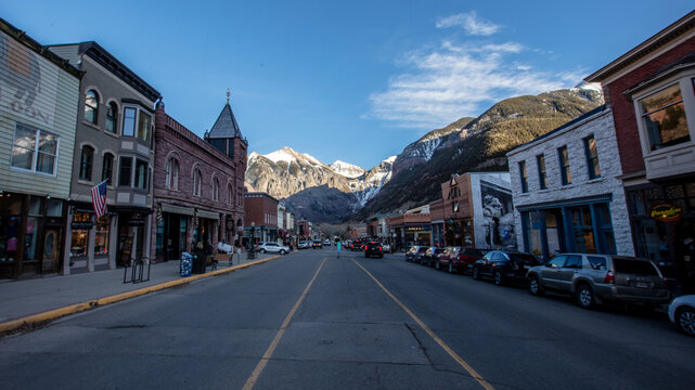 Telluride, Colorado, United States - 18 November 2021: View of the historic main street with classic brick buildings, parked cars, and snow-capped mountains under a clear blue sky.