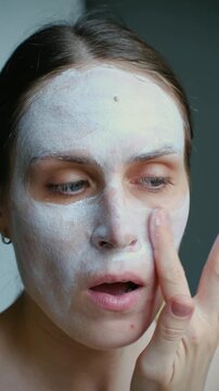 Young caucasian woman applying a cosmetic white facial mask to her skin, engaging in a daily beauty routine for skin treatment and rejuvenation in the comfort of her home bathroom. Vertical video.
