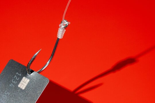 Credit card caught on a silver fishing hook against a red background with copy space. Financial fraud, cyber security, phishing scams, and debt traps concept.