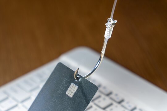 Fishing hook piercing a credit card over a blurred laptop keyboard. Online phishing scams, cybersecurity threats, digital fraud, and internet identity theft concept.