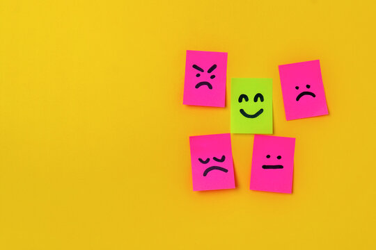 Green sticky note with a happy face surrounded by pink notes displaying negative expressions on a yellow background with copy space. Mental health concept.