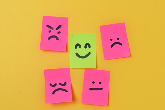 Green sticky note with a happy face surrounded by pink notes displaying negative expressions on a yellow background with copy space. Mental health concept.
