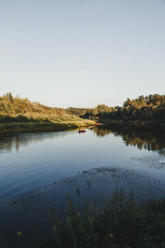 Cesis, Cesis Municipality, Latvia - 26 July 2023: View of a red canoe paddling on the calm Gauja River surrounded by lush green forests and reflections under a clear sky.