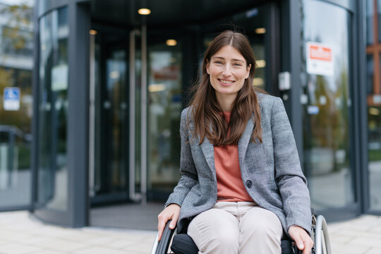 Woman smiles warmly from wheelchair near building entrance. She wears a gray coat over a rust-colored top. Her long brown hair falls gently over her shoulders