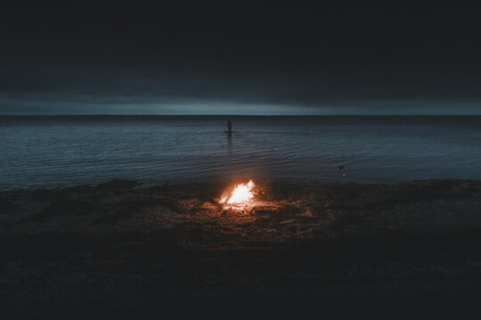 Laane County, Estonia - 26 August 2023: View of a bonfire burning on the rocky shore of Vormsi at night, with a lone person standing in the calm sea under a dark sky.