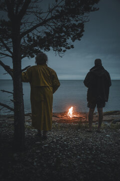Laane County, Estonia - 26 August 2023: View of two people standing by a small bonfire on a rocky beach at dusk with the sea in the background and a tree in the foreground.