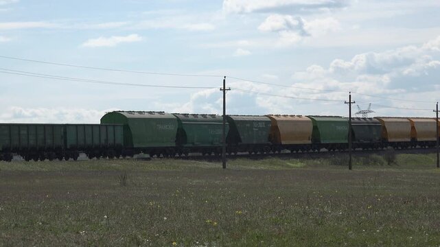 Mykolaiv, Ukraine &ndash; Apr 30, 2020: Long freight train with grain hopper and gondola wagons crossing flat open country past a spring meadow, under bright cumulus clouds