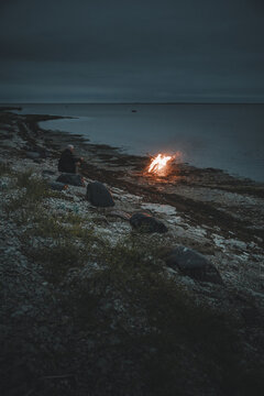 Laane County, Estonia - 26 August 2023: View of a person sitting by a campfire on the rocky shore of Vormsi at dusk, with the dark sea and evening sky in the background.