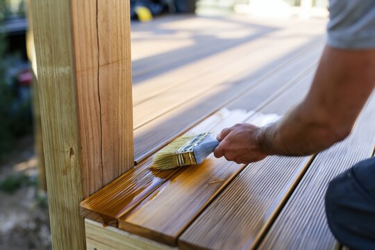 Hand applying oil to a wooden deck with a flat brush, showing a wet sheen on caramel brown planks for home improvement and DIY maintenance