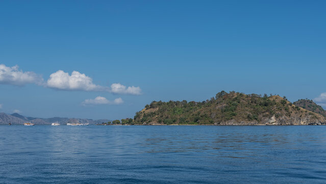 An island in the ocean. There is green vegetation on the slopes. Tourist ships are not far from the coast. Blue sky, clouds. Indonesia. Kelor Island 