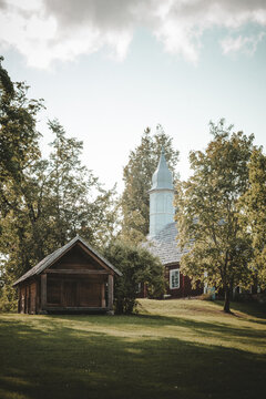 Sigulda, Latvia - 25 July 2023: View of the historic Sigulda Lutheran Church with its distinctive spire and a traditional wooden outbuilding set amidst lush green trees under a cloudy sky.