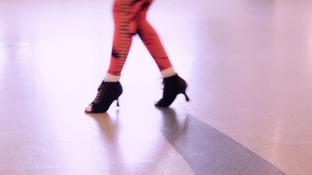 Rehearsal step. Dance instructor. Female legs in bright leggings energizing moving on black high heels on light studio floor background.