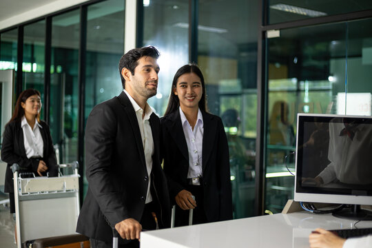 Business travelers arriving at modern office reception with luggage, smiling colleagues in formal suits demonstrating teamwork, travel, hospitality and professional welcome