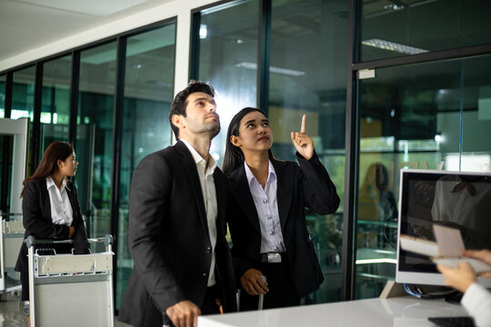 Business travelers at office reception with luggage as woman points upward and man looks above, showing guidance, direction, teamwork, travel, workplace and professional assistance