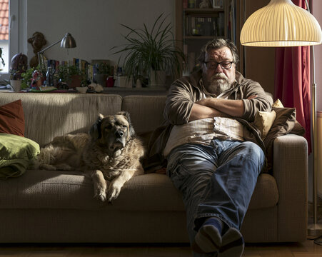A middle-aged man with glasses and a beard sits on a sofa with his arms crossed, looking tired and bored. Beside him, a large dog with brown fur rests its head on the sofa cushion.