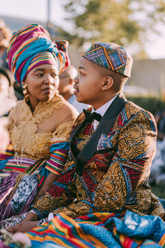 A vibrant African wedding ceremony with a couple dressed in colorful traditional attire. The woman wears a striking headwrap and ornate dress, while the man sports a patterned suit and matching cap.