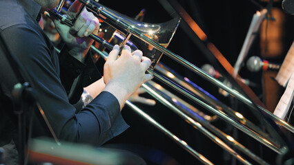 Closeup of musician playing trombone during symphony orchestra performance. Hands grip slide with precision under stage lighting. Polished brass reflects colorful spotlights. Dark formal attire © VeNN