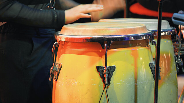 Closeup of glossy conga drums in warm yellow orange gradient under stage lighting. Musician hand visible mid motion. Dynamic composition highlights percussion texture performance energy