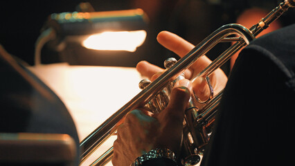 Hands gripping brass trumpet under warm stage light during symphony orchestra rehearsal. Shallow depth of field highlights instrument fingers. Dark background with soft bokeh © VeNN