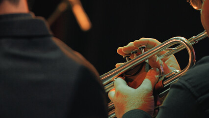 Closeup of musician hands playing trumpet under warm stage lights during symphony orchestra concert. Dark background highlights instrument fingers on valves. Evokes focus, precision, musical passion © VeNN