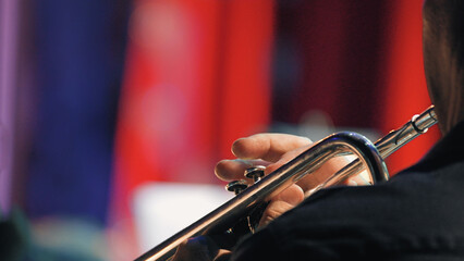 Closeup of musician playing trumpet during symphony orchestra concert. Hands on instrument, dark attire, vibrant red blue stage lights create dramatic mood. Ideal for music, performance, arts events © VeNN
