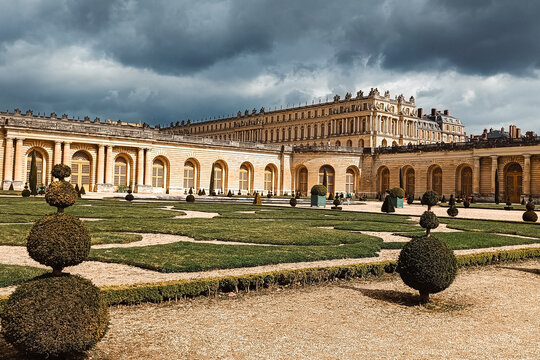 Versailles, France - 02 April 2026: View of the Palace of Versailles with its manicured gardens, topiary shrubs, and historic architecture under a dramatic cloudy sky.