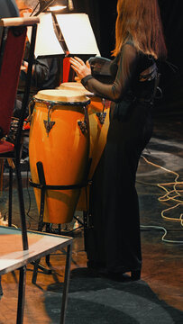 Female percussionist in black outfit playing conga drums under stage lights during symphony rehearsal. Warm orange drums contrast with dark stage