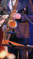 Closeup of musician playing saxophone during live orchestra performance. Warm golden tones of instrument contrast with cool blue stage lighting. Formal suit, microphone, music stand visible © VeNN
