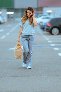 Contented Young Woman Posing In Supermarket Parking Lot Holding Grocery Bag. Confident smiling female customer stands with paper shopping bag outdoors near car park