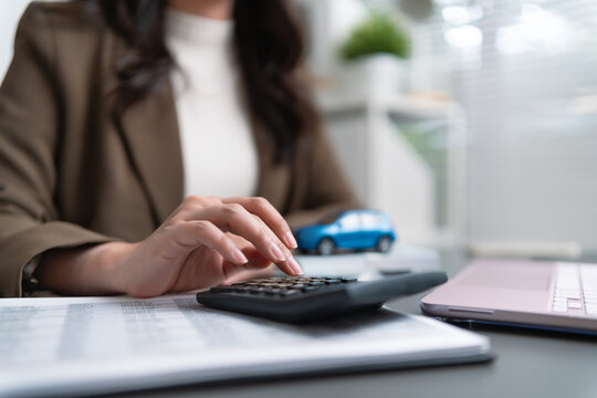 Businesswoman using a calculator beside a car model and laptop, representing car insurance, auto loan payments, vehicle financing, purchase costs, budgeting, financial planning, and ownership.