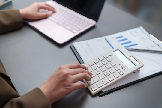 Businesswoman using a calculator and laptop to review financial reports with charts and data, representing accounting, budgeting, expense calculation, business analysis, planning, and finance.