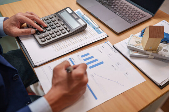 Businessman using a calculator to analyze financial reports beside cash, house and car models, representing mortgage, property investment, loan payments