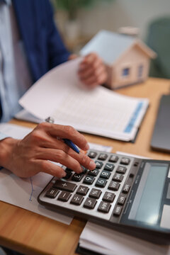 Businessman using a calculator with financial documents and a house model, representing mortgage calculation, home loan payments, property finance, housing costs, budgeting, investment, and ownership.