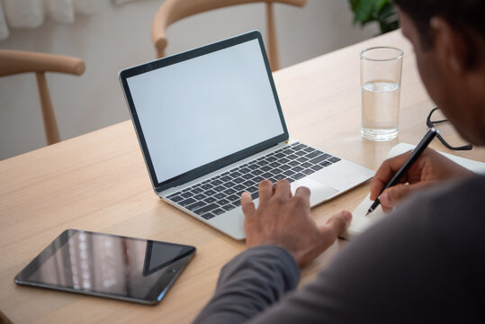 African Man working remotely on a laptop while writing notes at a home desk with a tablet, representing online business, planning, productivity