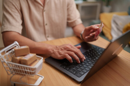 Man using a laptop and credit card for online shopping at home, with a miniature shopping cart and parcels representing secure digital payment, e-commerce, retail purchase, and delivery.
