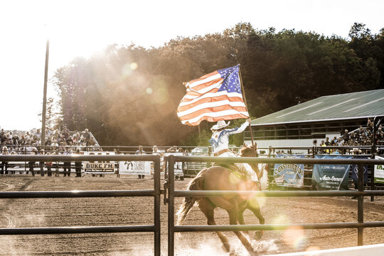 Saint Johns, Michigan, United States - 12 August 2017: View of a rodeo rider on horseback carrying an American flag in an arena during the golden hour with spectators in the background.