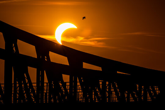 Partial solar eclipse above a large industrial bridge structure, creating a dramatic silhouette against a glowing orange sky at sunset.