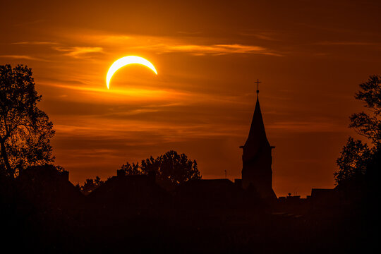 Partial solar eclipse above a church and silhouetted trees against a dramatic orange sunset sky, creating a striking celestial and atmospheric landscape.