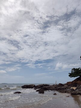 An eye-level, wide perspective looking across barnacle-covered rocks on a sandy shoreline with gentle sea waves washing past, under a bright, partly cloudy sky.
